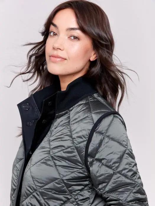 A person with long hair wears a black Charlie B Reversible Quilted Puffer Jacket with a button-up front, a black sweater featuring a white design, and light-colored pants, creating a versatile style perfect for any winter wardrobe. Standing against a plain background, their look is both stylish and adaptable.