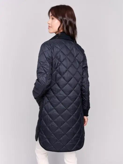 A person with long hair wears a black Charlie B Reversible Quilted Puffer Jacket with a button-up front, a black sweater featuring a white design, and light-colored pants, creating a versatile style perfect for any winter wardrobe. Standing against a plain background, their look is both stylish and adaptable.