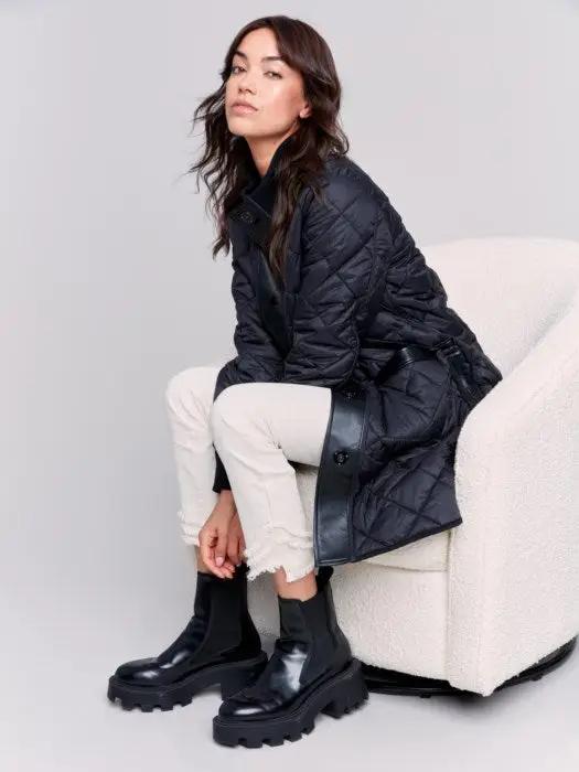 A person with long hair wears a black Charlie B Reversible Quilted Puffer Jacket with a button-up front, a black sweater featuring a white design, and light-colored pants, creating a versatile style perfect for any winter wardrobe. Standing against a plain background, their look is both stylish and adaptable.