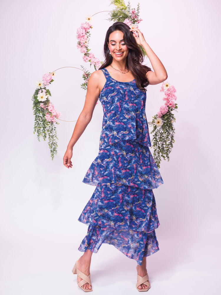 A woman gracefully poses in front of floral decorations wearing the Elena Wang Tiered Round Neck Dress in Blue and beige heels.