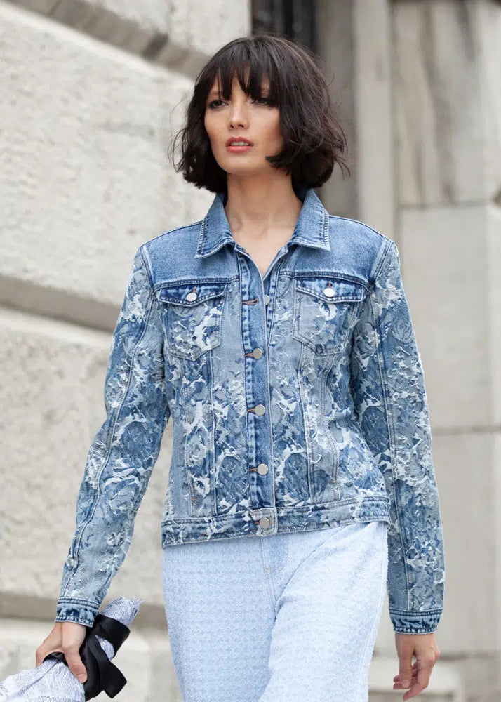 A stylish person with short brown hair is wearing an Elena Wang Floral Denim Jacket and light blue pants, walking outdoors near a stone wall.