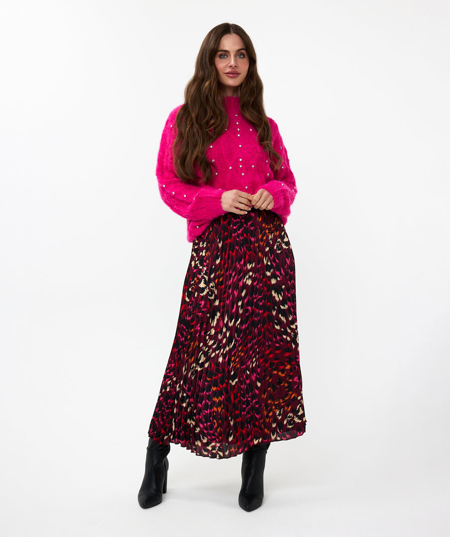 A person stands against a white background, wearing a high-quality bright pink sweater with the Esqualo Pleated Pink Floral Skirt, paired with black ankle boots.