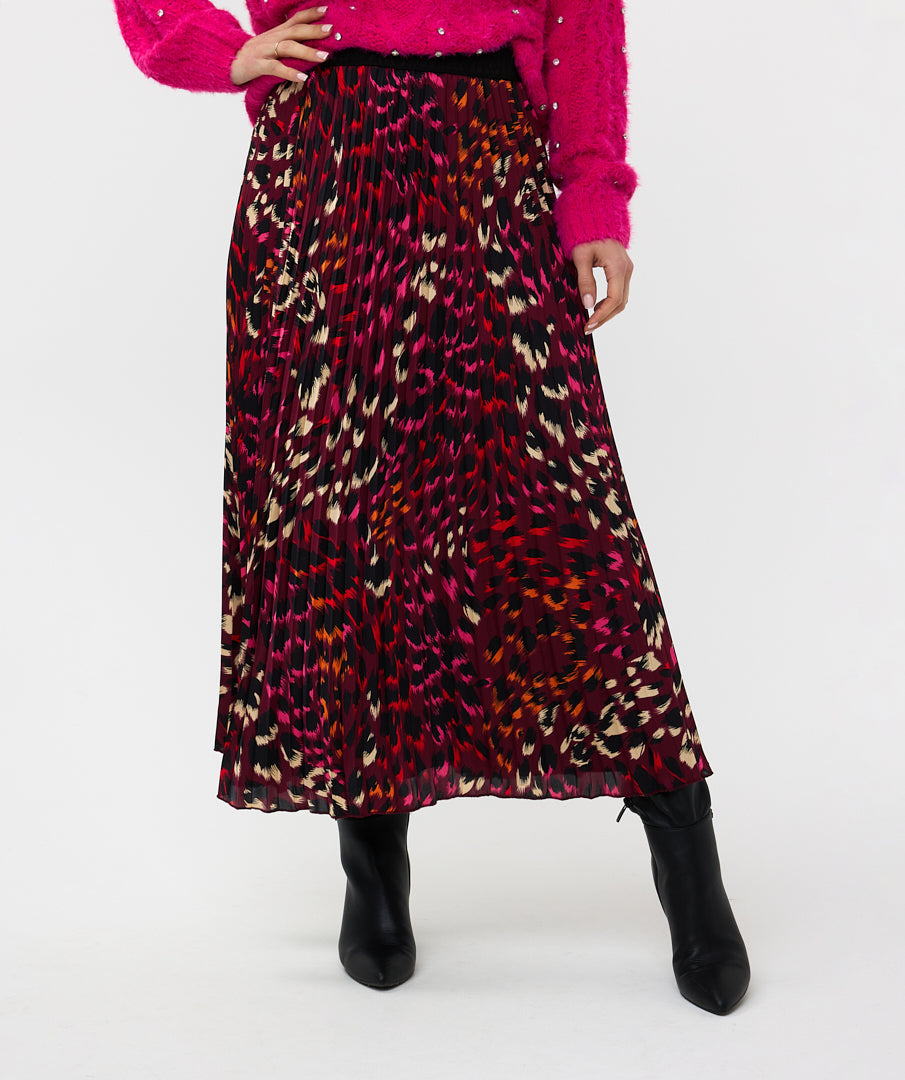 A person stands against a white background, wearing a high-quality bright pink sweater with the Esqualo Pleated Pink Floral Skirt, paired with black ankle boots.