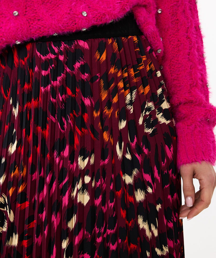 A person stands against a white background, wearing a high-quality bright pink sweater with the Esqualo Pleated Pink Floral Skirt, paired with black ankle boots.