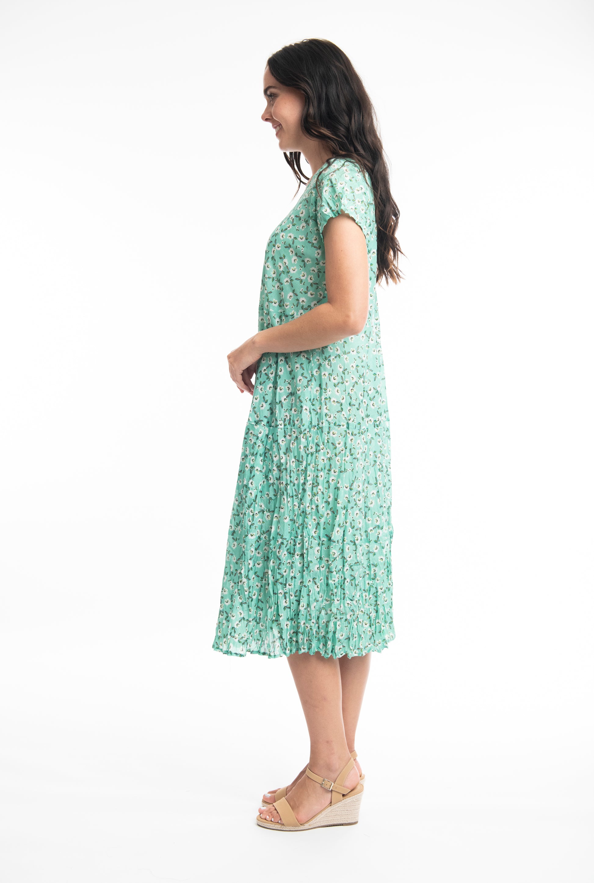 A woman in a Orientique Sweet Bias Green Dress smiling and posing with one hand on her hip against a white background.