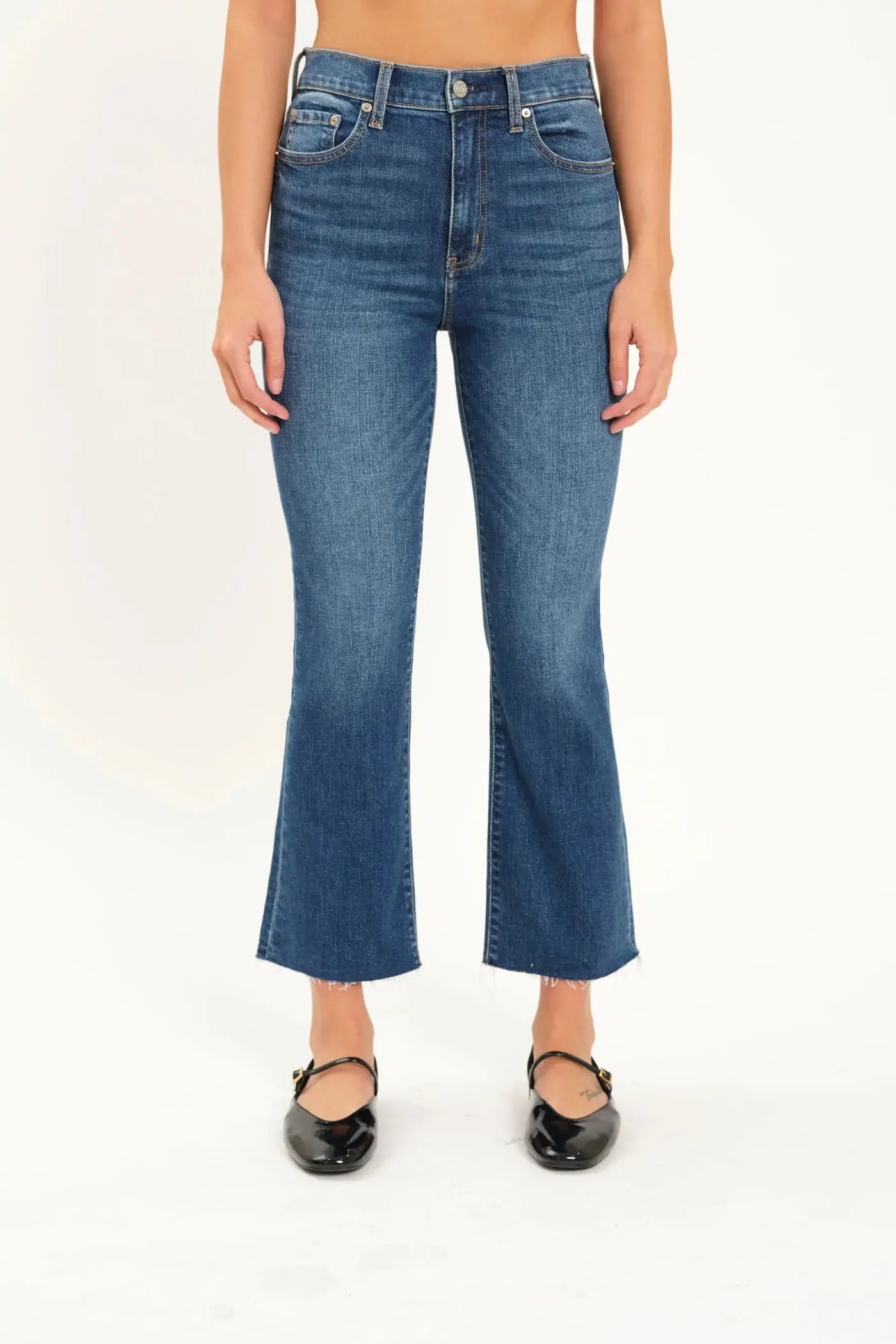 A person wearing Daze's Shy Girl High Rise Crop Flare jeans in denim blue and black ballet flats is standing against a plain white background.