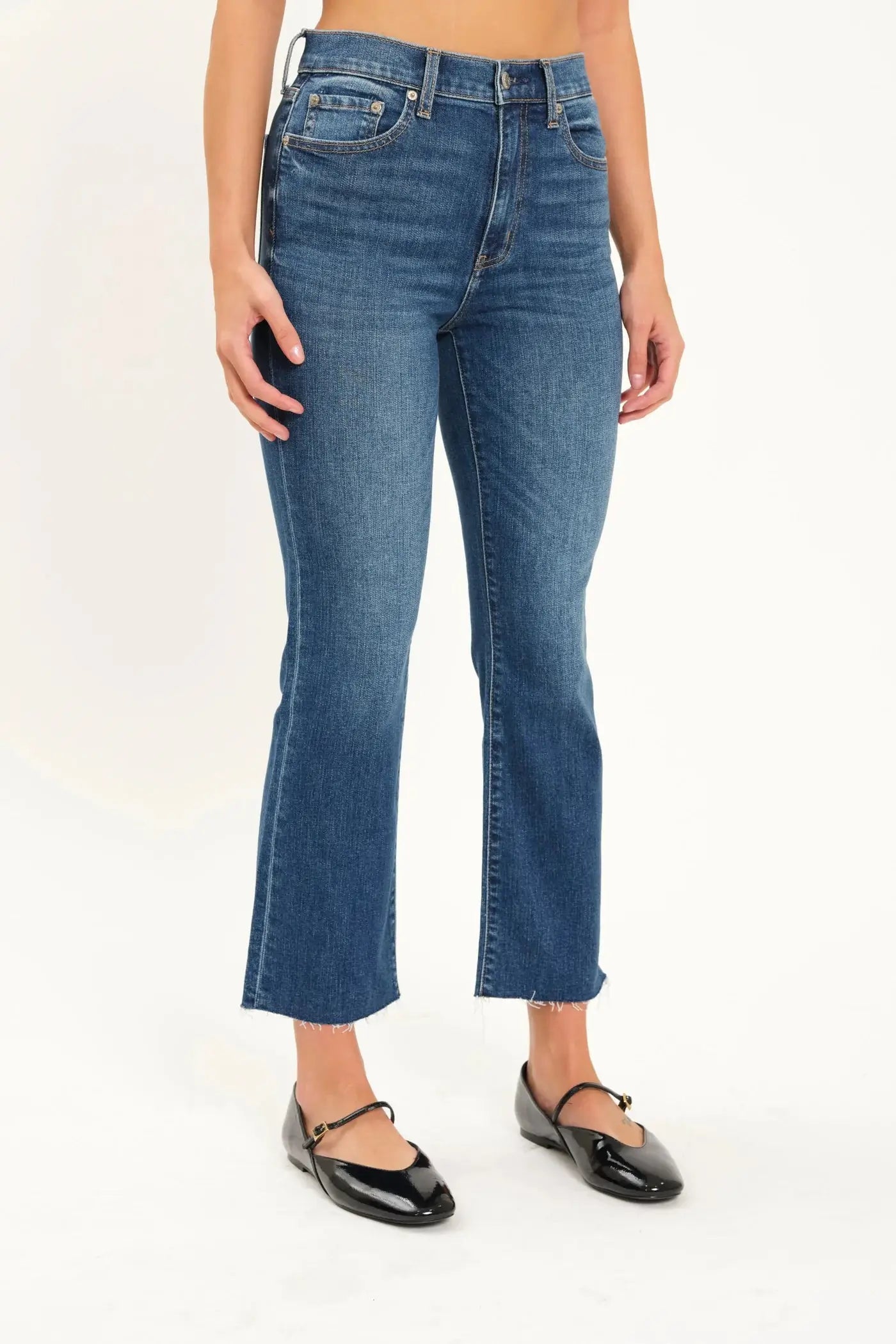 A person wearing Daze's Shy Girl High Rise Crop Flare jeans in denim blue and black ballet flats is standing against a plain white background.