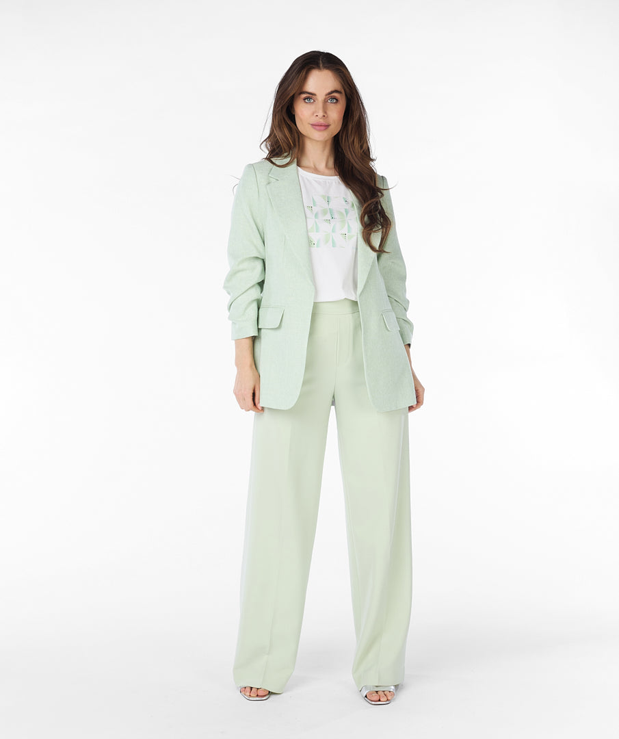 A woman with long brown hair wears a high-quality linen light green Esqualo Pastel Blazer over a white patterned t-shirt and light green pants, standing against a plain white background. The blazer serves as a versatile piece suitable for various occasions.