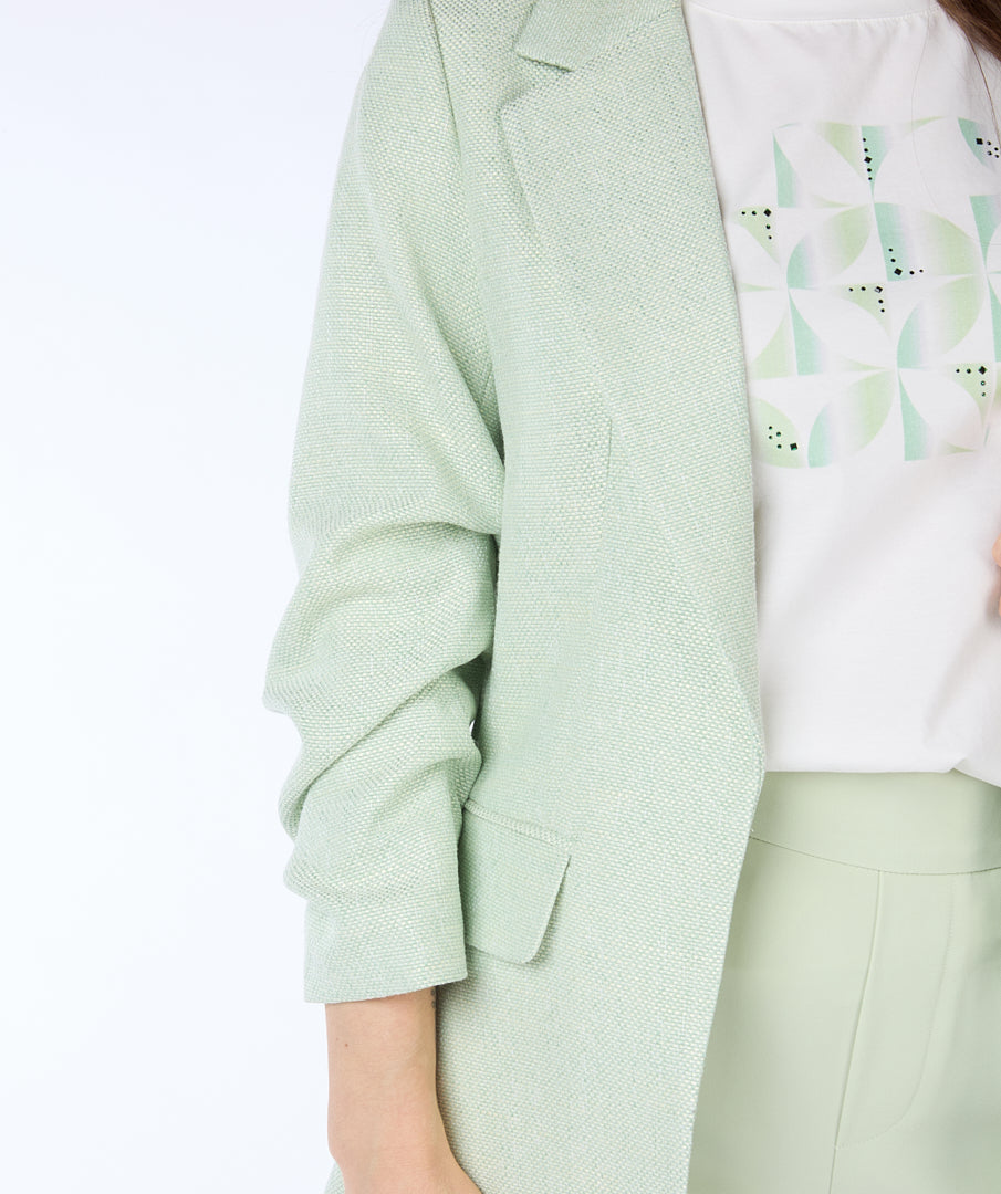 A woman with long brown hair wears a high-quality linen light green Esqualo Pastel Blazer over a white patterned t-shirt and light green pants, standing against a plain white background. The blazer serves as a versatile piece suitable for various occasions.