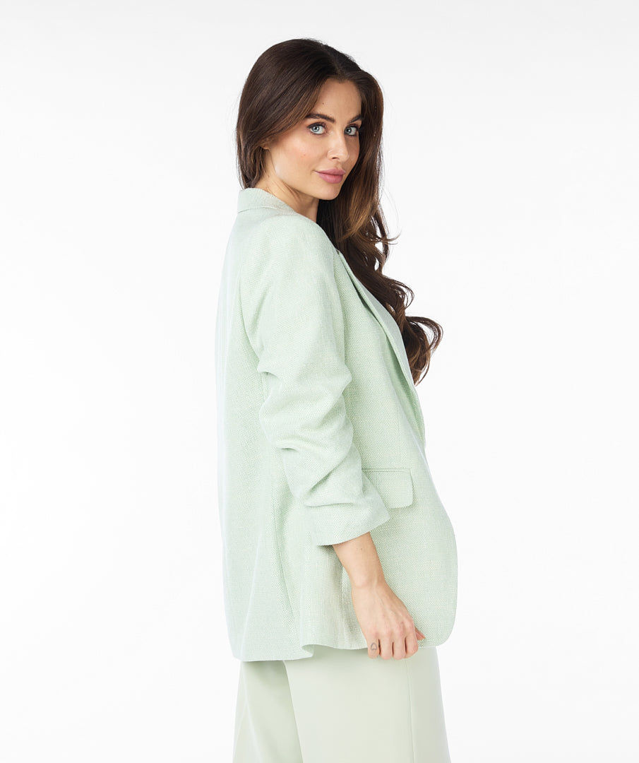 A woman with long brown hair wears a high-quality linen light green Esqualo Pastel Blazer over a white patterned t-shirt and light green pants, standing against a plain white background. The blazer serves as a versatile piece suitable for various occasions.