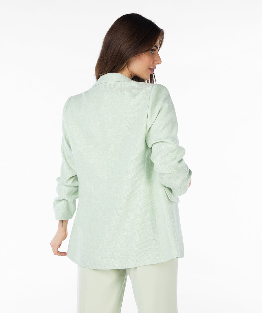 A woman with long brown hair wears a high-quality linen light green Esqualo Pastel Blazer over a white patterned t-shirt and light green pants, standing against a plain white background. The blazer serves as a versatile piece suitable for various occasions.