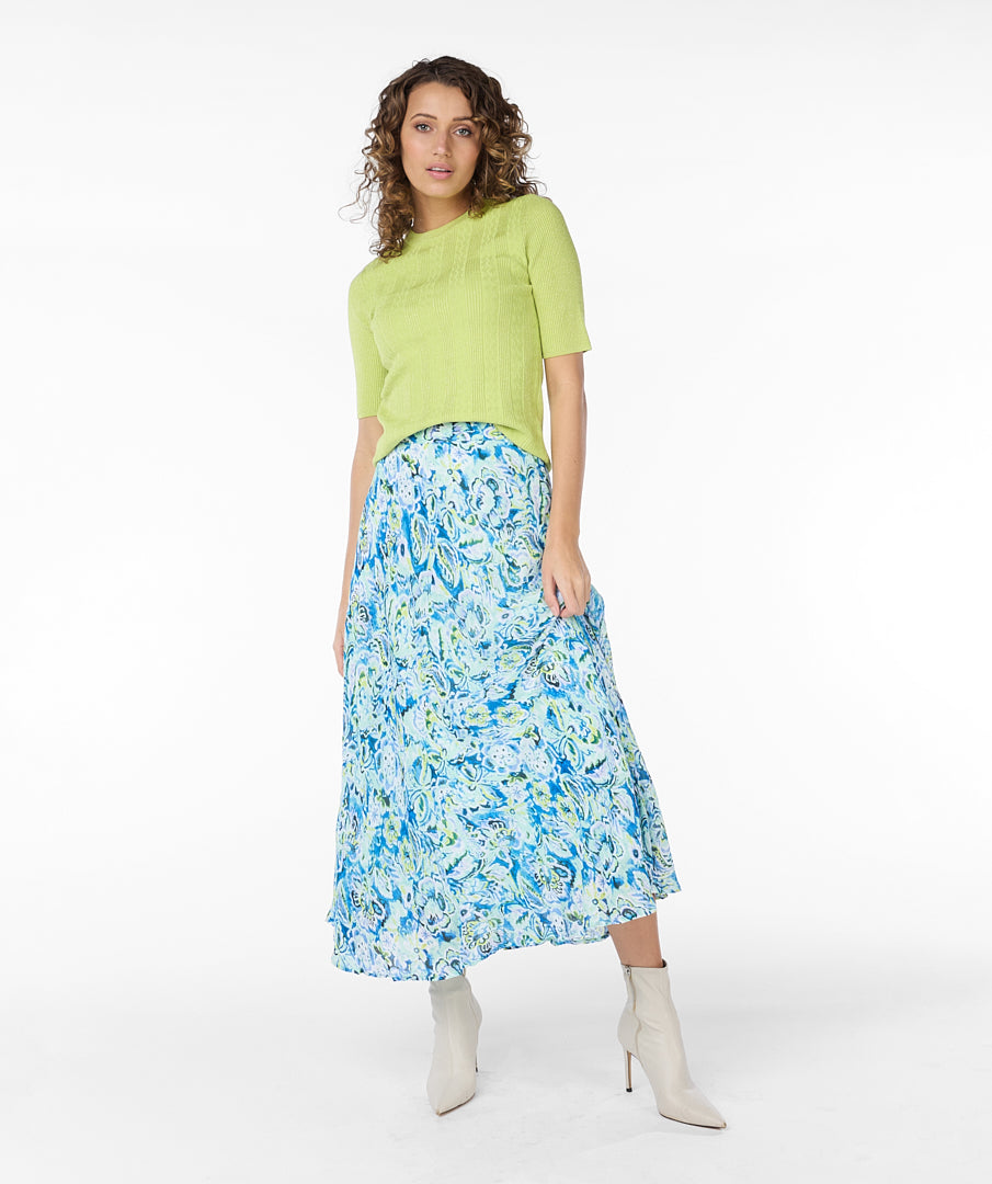 A modern woman with curly hair stands against a plain white background, embodying timeless elegance in a lime green short-sleeved top, white ankle boots, and a vibrant blue and white Esqualo Bayside Skirt adorned with floral patterns.