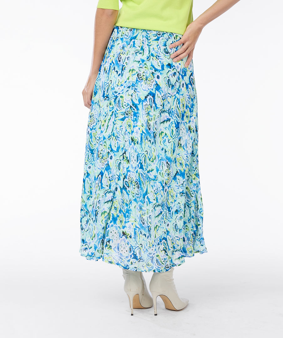 A modern woman with curly hair stands against a plain white background, embodying timeless elegance in a lime green short-sleeved top, white ankle boots, and a vibrant blue and white Esqualo Bayside Skirt adorned with floral patterns.