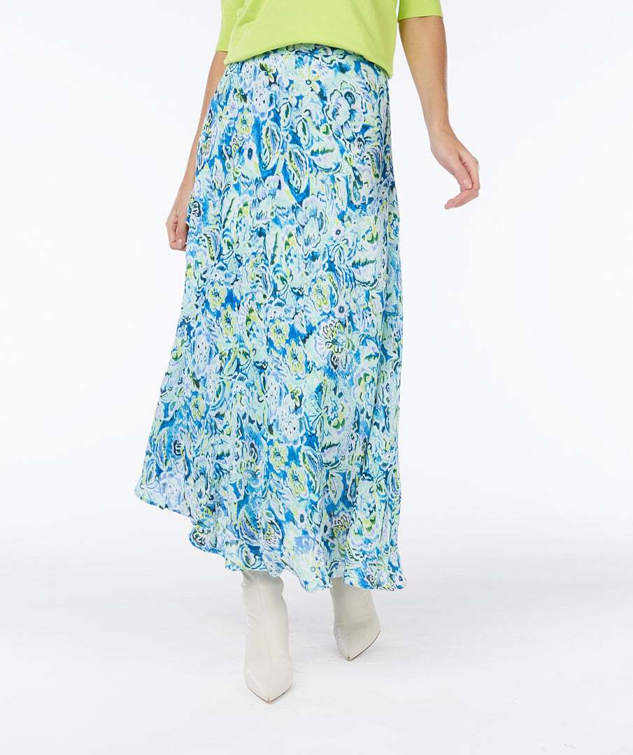 A modern woman with curly hair stands against a plain white background, embodying timeless elegance in a lime green short-sleeved top, white ankle boots, and a vibrant blue and white Esqualo Bayside Skirt adorned with floral patterns.