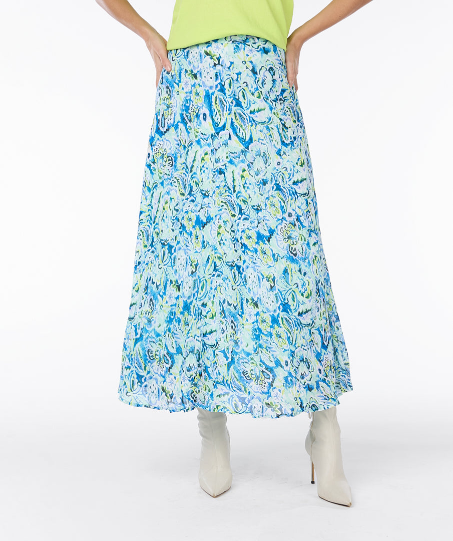 A modern woman with curly hair stands against a plain white background, embodying timeless elegance in a lime green short-sleeved top, white ankle boots, and a vibrant blue and white Esqualo Bayside Skirt adorned with floral patterns.