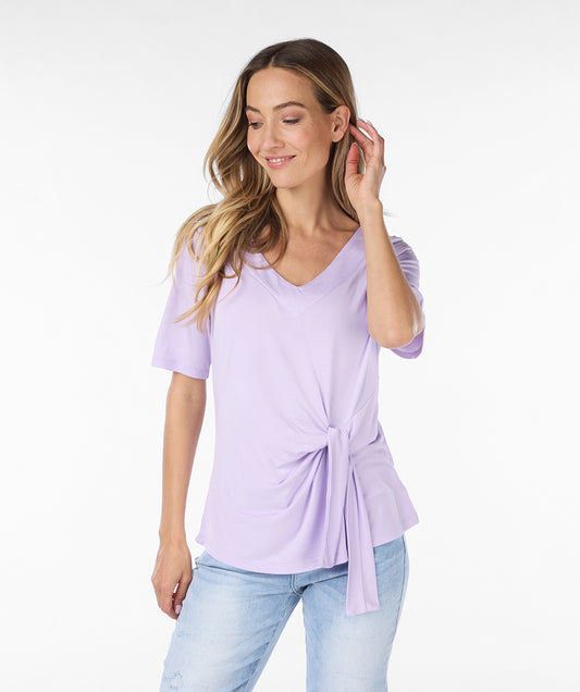 A woman with long blonde hair is standing and looking over her shoulder. She is wearing an adorable and comfortable Lilac Top by Esqualo, paired with light blue jeans against a plain white background.