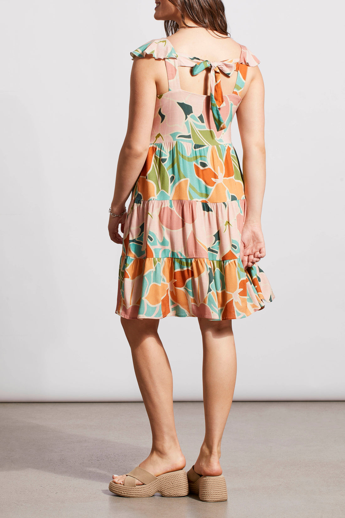 A woman in a Tribal sleeveless dress with lining, floral print and tie back, paired with beige sandals standing against a plain backdrop.