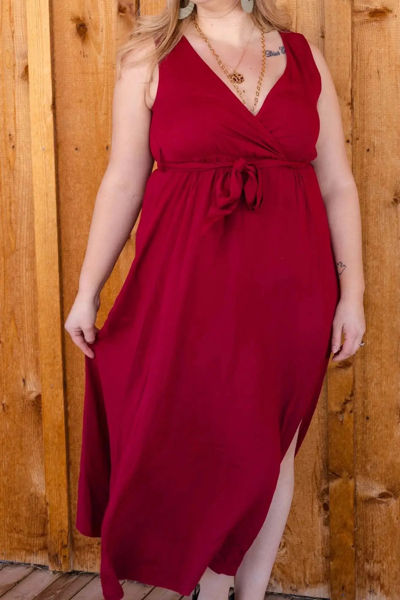 Woman in a wine red V Neck Cross-Tie Sleeve dress from Don't Be Chy Boutique posing between wooden beams, with visible chunky gold necklace and beige heels.