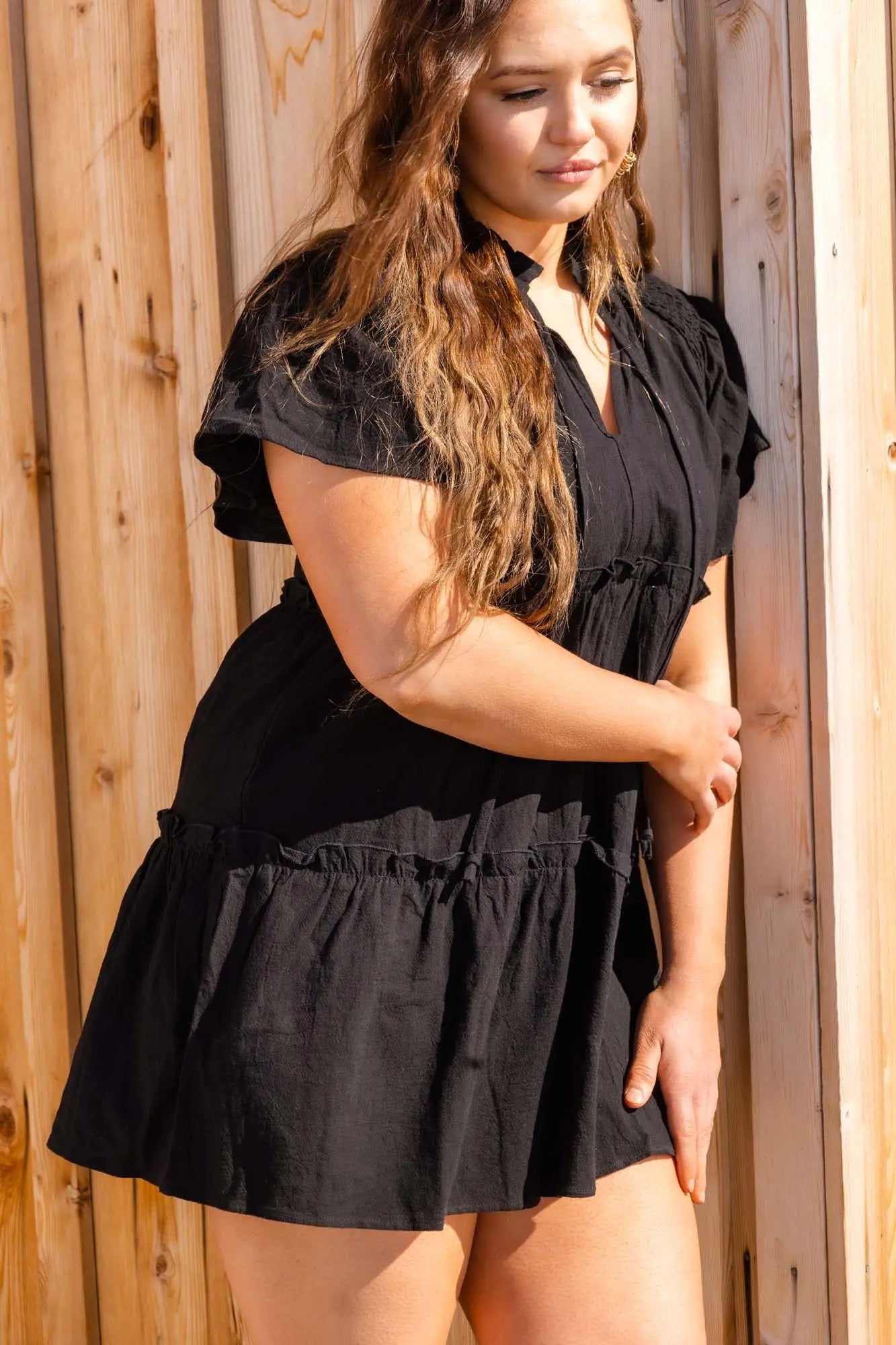 A woman in a black ruffled mini dress with puff sleeves and a tied neckline, holding the skirt, standing outdoors by Don't Be Chy Boutique.