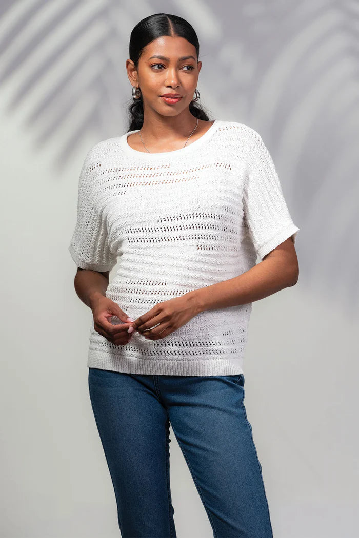 A woman in a white Round Neck Knit Sweater by Alison Sheri and blue jeans stands with her hands together, set against a light backdrop with shadow patterns.