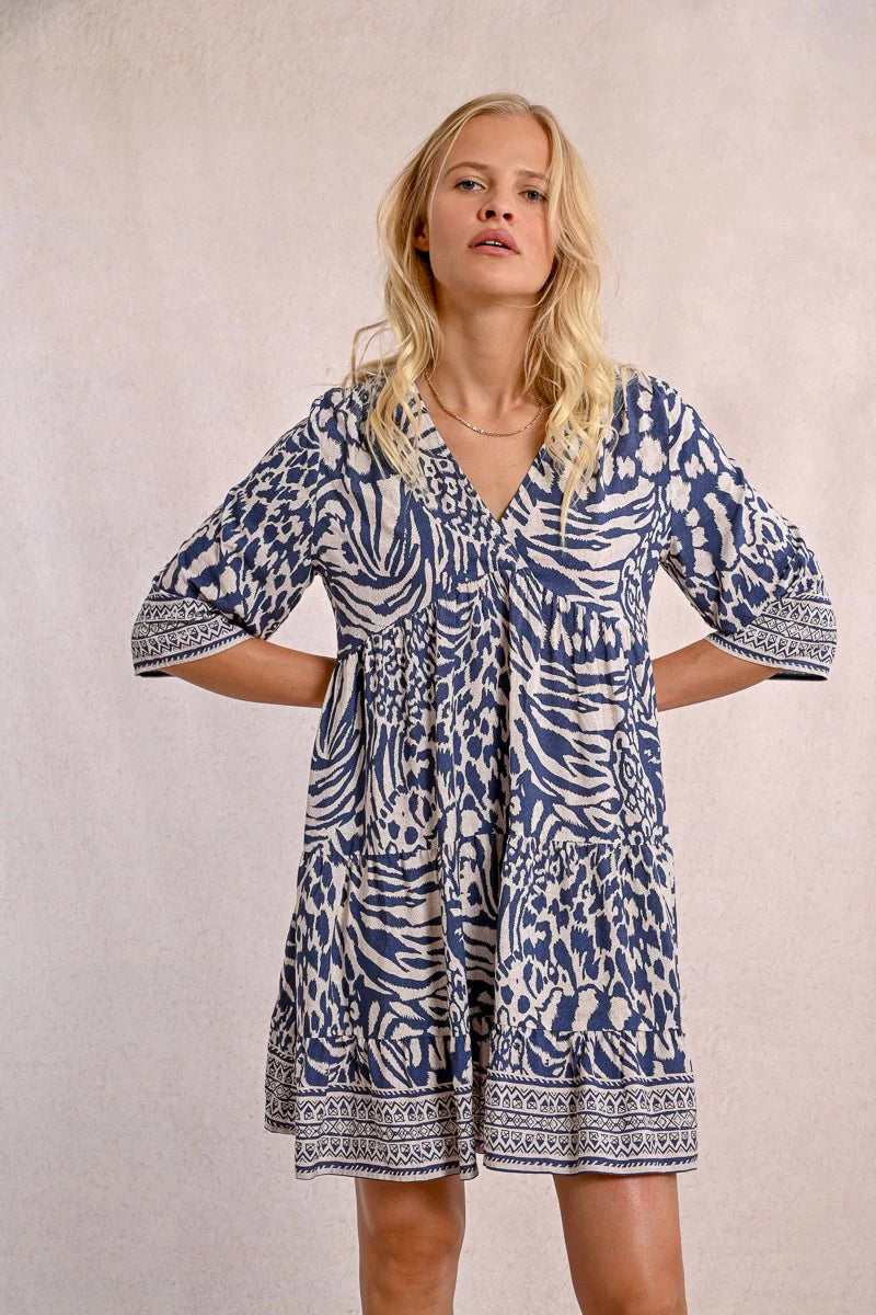 A person with long blonde hair stands against a plain backdrop, wearing the Molly Bracken 3/4 Sleeve Kimono Dress in a blue zebra print and sandals, looking to the right.