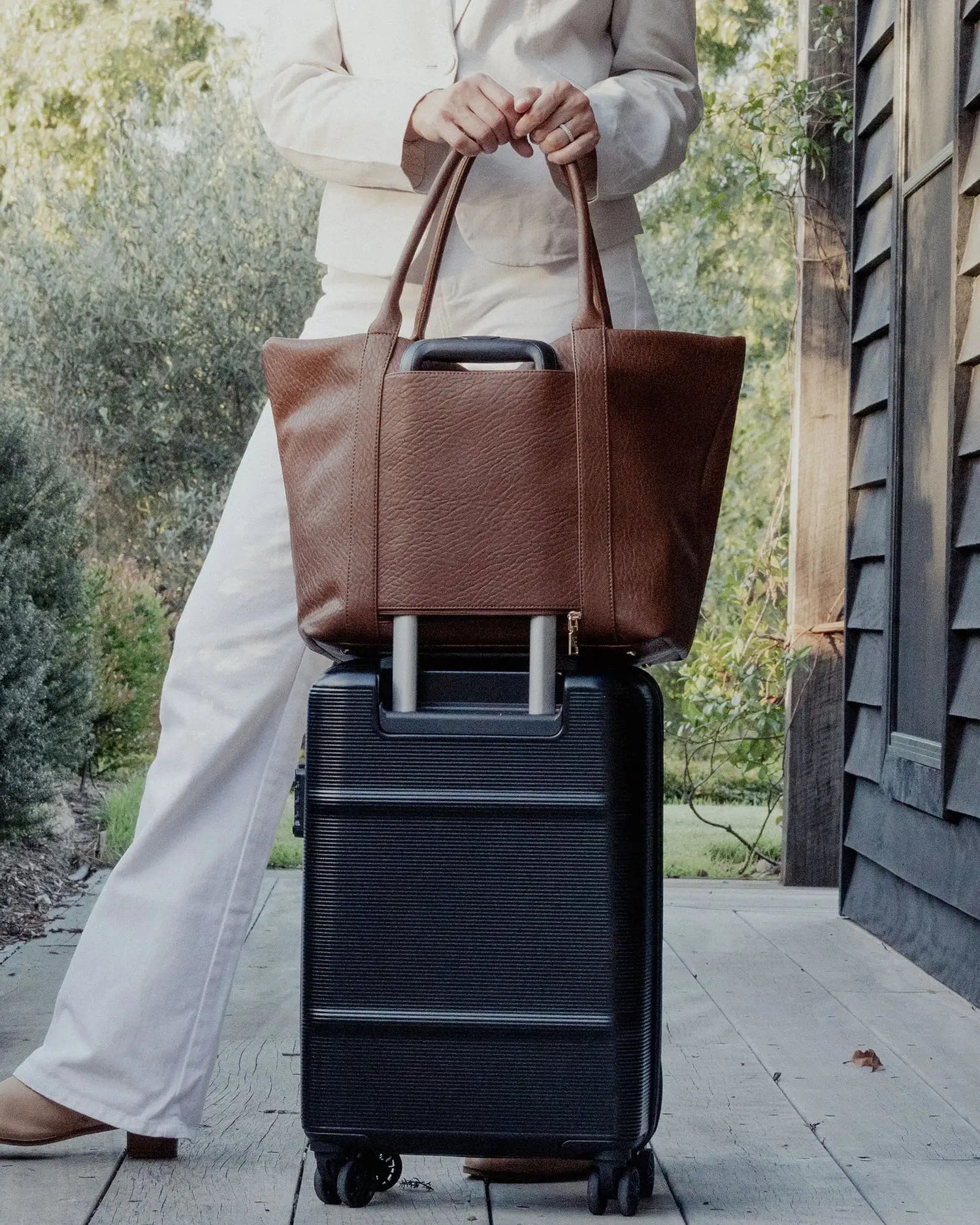 A person in white clothing stands on a porch holding The Savannah Tote Bag by Louenhide, crafted from vegan leather, resting it atop a black rolling suitcase.