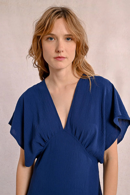 A person elegantly poses against a plain backdrop in a blue Front Pleat Long Marine Dress by Molly Bracken, characterized by its flowy design and short sleeves.