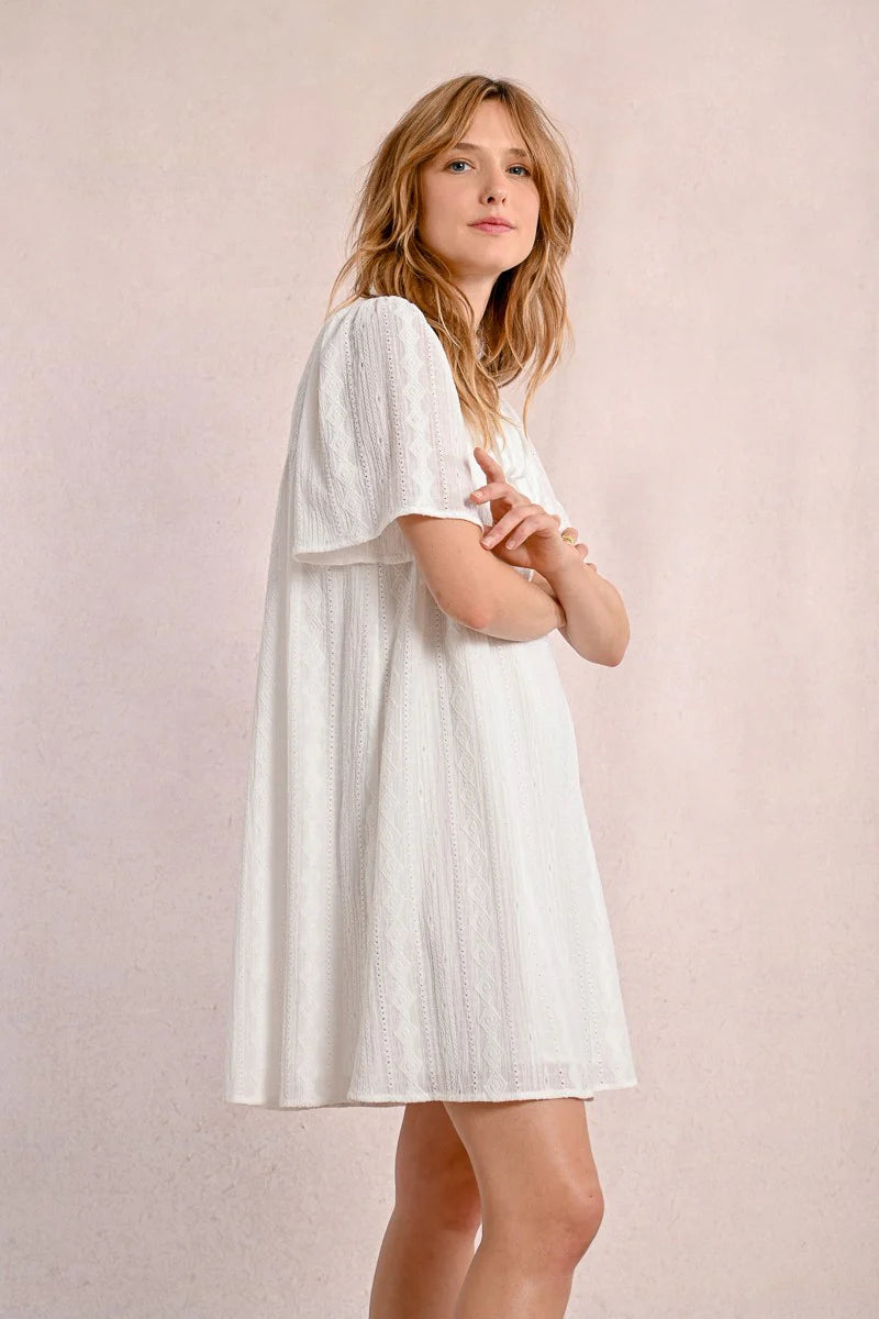 Someone in a Molly Bracken Off White Woven Dress with a v-neck and brown sandals stands against a plain background, looking to the side.