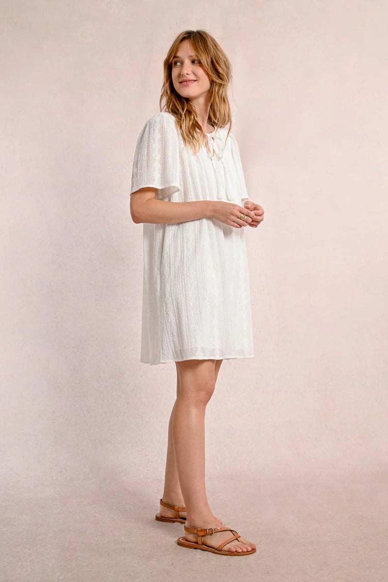 Someone in a Molly Bracken Off White Woven Dress with a v-neck and brown sandals stands against a plain background, looking to the side.