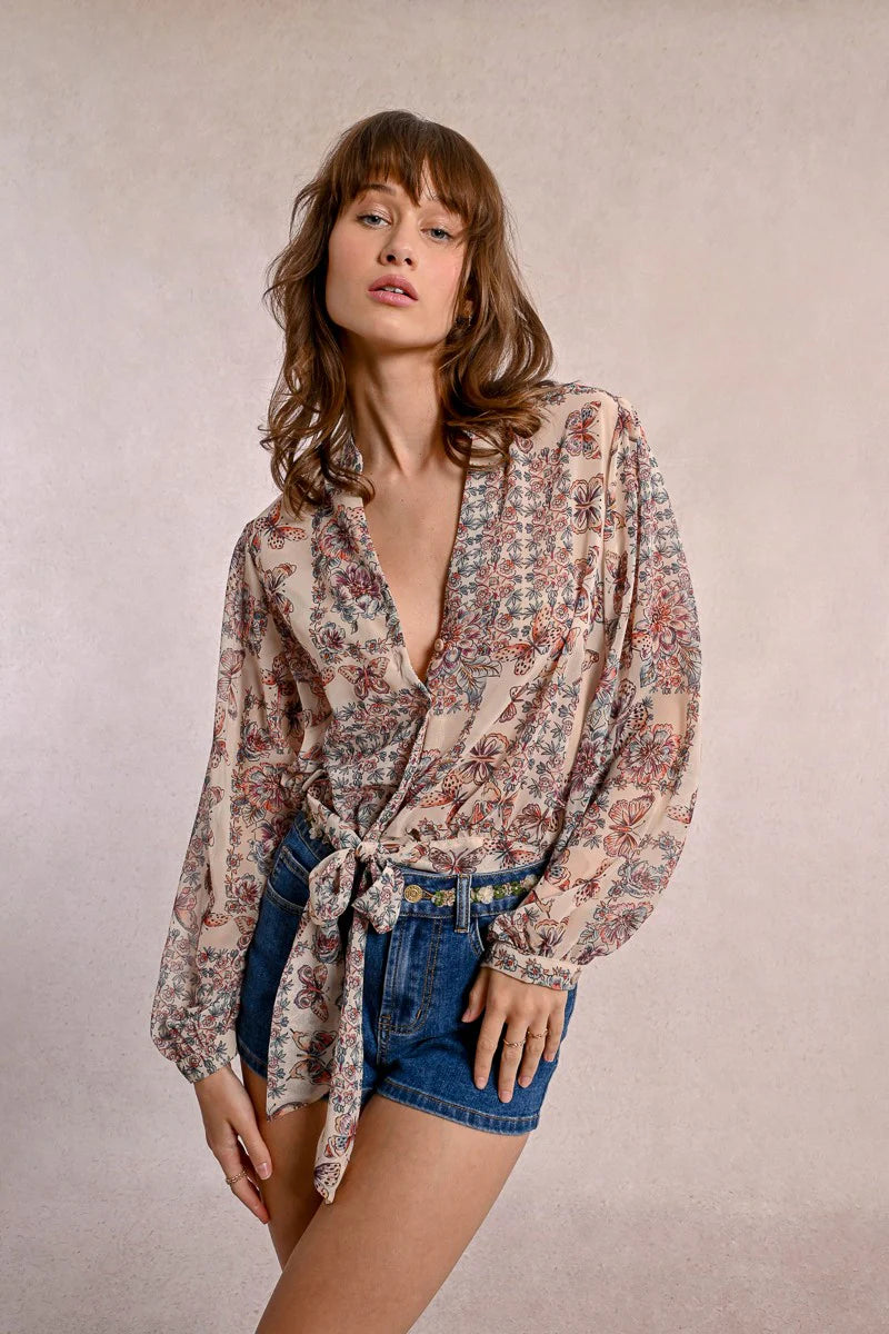 A person in a semi-transparent floral blouse and denim shorts stands against a plain background, elegantly showcasing the Front Tie Printed Blouse by Molly Bracken.