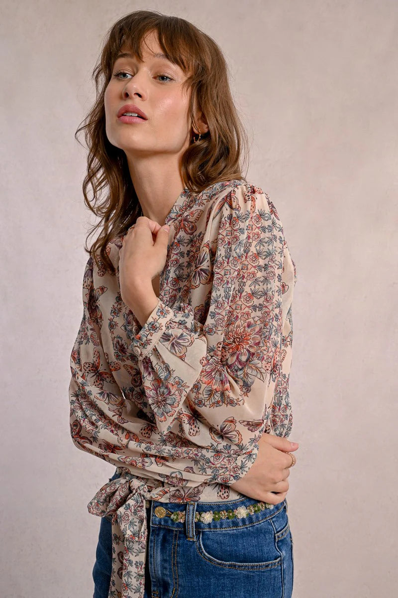 A person in a semi-transparent floral blouse and denim shorts stands against a plain background, elegantly showcasing the Front Tie Printed Blouse by Molly Bracken.