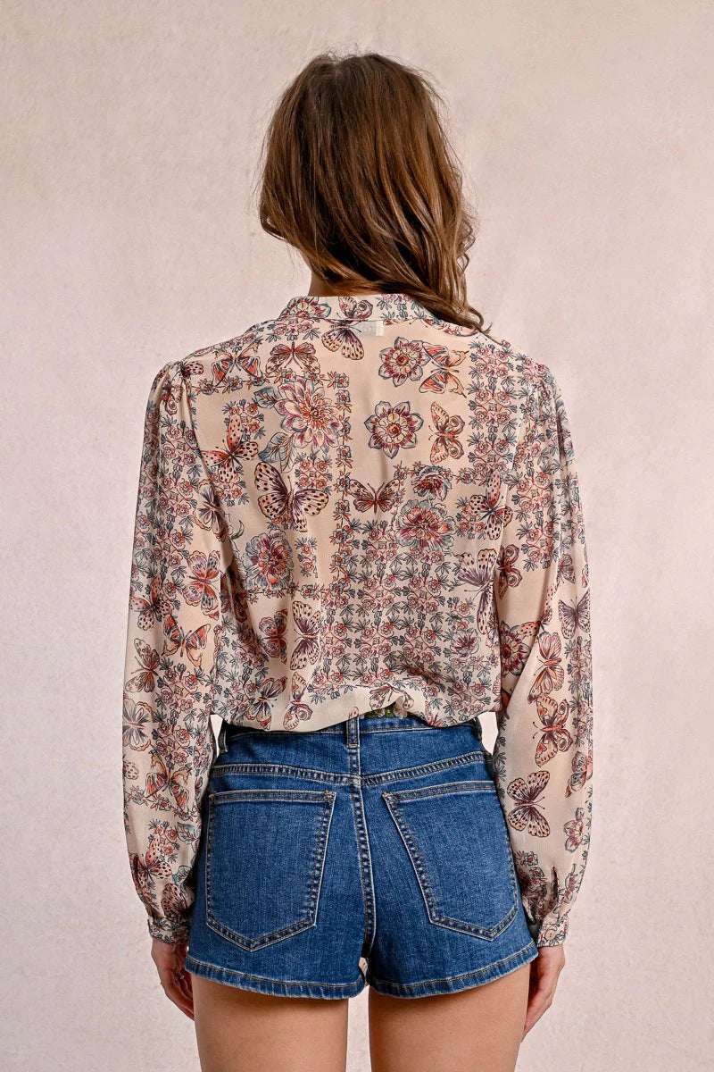 A person in a semi-transparent floral blouse and denim shorts stands against a plain background, elegantly showcasing the Front Tie Printed Blouse by Molly Bracken.