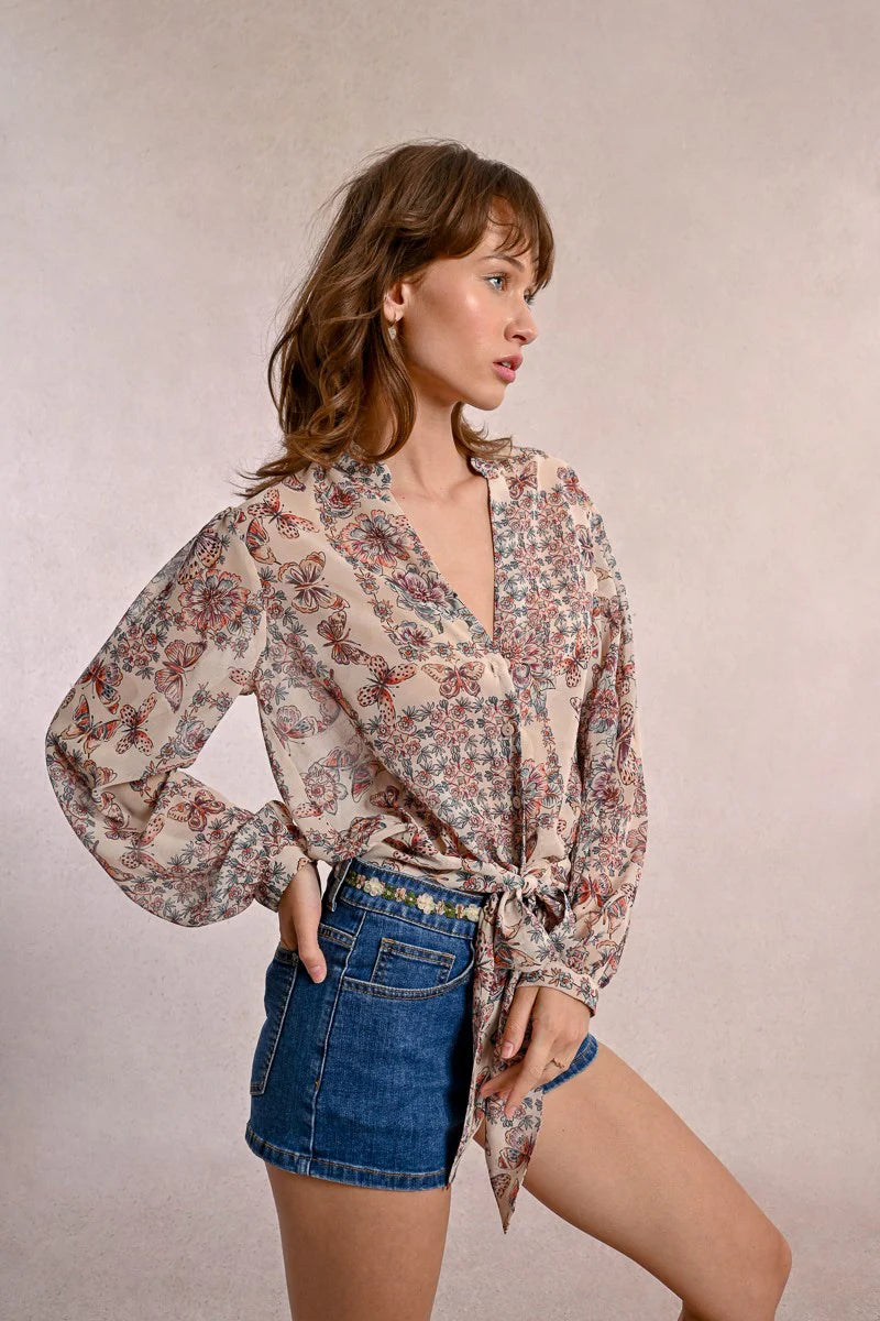 A person in a semi-transparent floral blouse and denim shorts stands against a plain background, elegantly showcasing the Front Tie Printed Blouse by Molly Bracken.