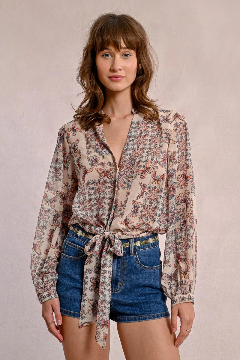 A person in a semi-transparent floral blouse and denim shorts stands against a plain background, elegantly showcasing the Front Tie Printed Blouse by Molly Bracken.