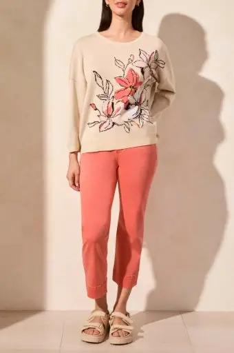 A person in a Tribal 3/4 Sleeve Crew Neck Sweater with a pink floral design, coral pants, and beige sandals stands against a light background.