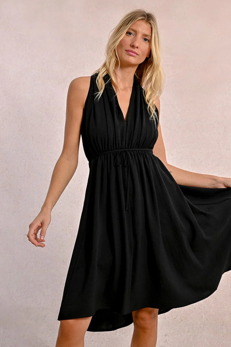 In a Short Pleated Dress by Molly Bracken, a woman exudes flirty elegance with its sleeveless design and plunge v-neck as she stands confidently against a plain background.