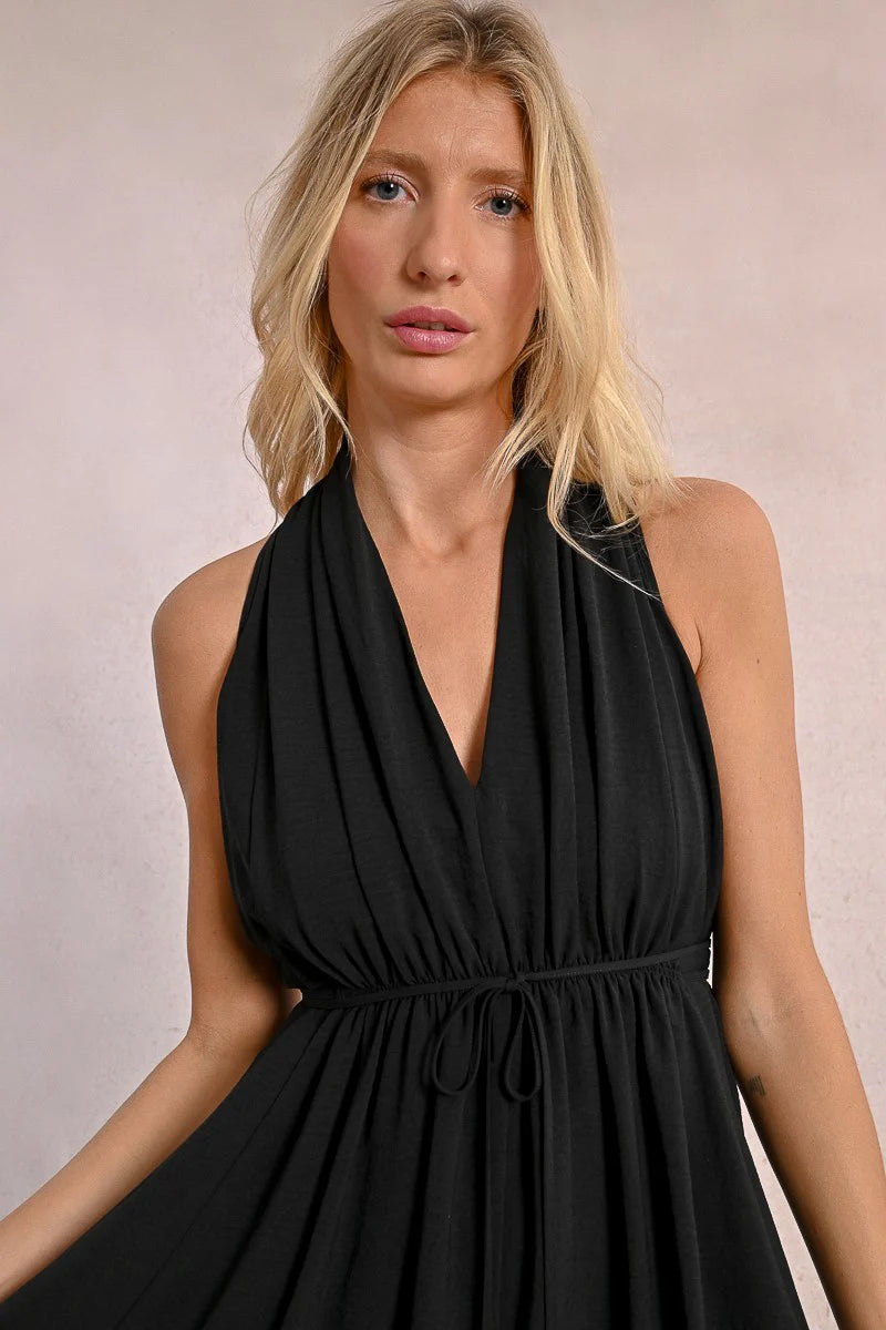 In a Short Pleated Dress by Molly Bracken, a woman exudes flirty elegance with its sleeveless design and plunge v-neck as she stands confidently against a plain background.