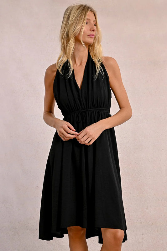 In a Short Pleated Dress by Molly Bracken, a woman exudes flirty elegance with its sleeveless design and plunge v-neck as she stands confidently against a plain background.