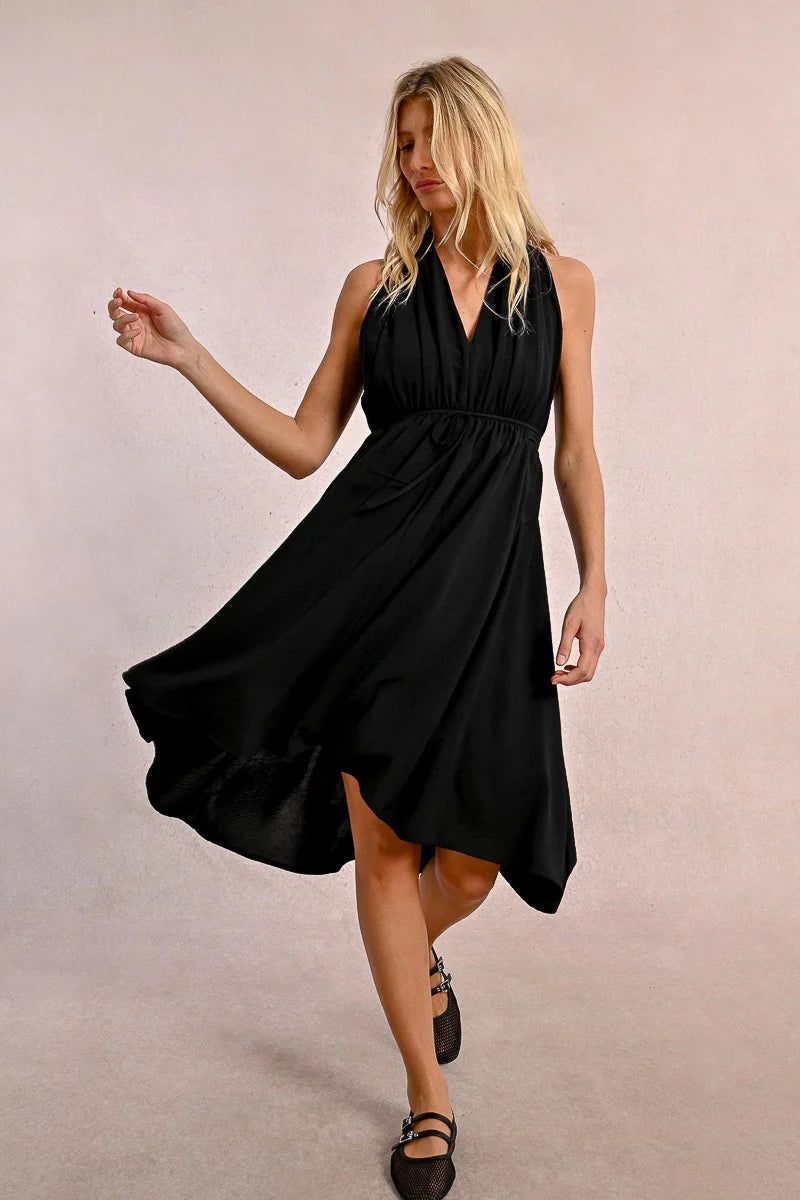 In a Short Pleated Dress by Molly Bracken, a woman exudes flirty elegance with its sleeveless design and plunge v-neck as she stands confidently against a plain background.