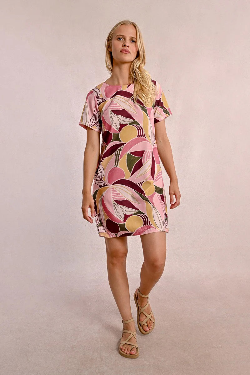 A woman in a Molly Bracken Floral Print T Shirt Dress, featuring a round collar, practical pockets, and short sleeves, stands against a neutral background. Her long blonde hair complements the vibrant outfit she pairs with chic sandals.