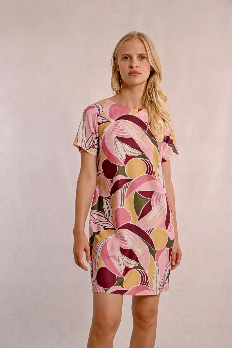 A woman in a Molly Bracken Floral Print T Shirt Dress, featuring a round collar, practical pockets, and short sleeves, stands against a neutral background. Her long blonde hair complements the vibrant outfit she pairs with chic sandals.