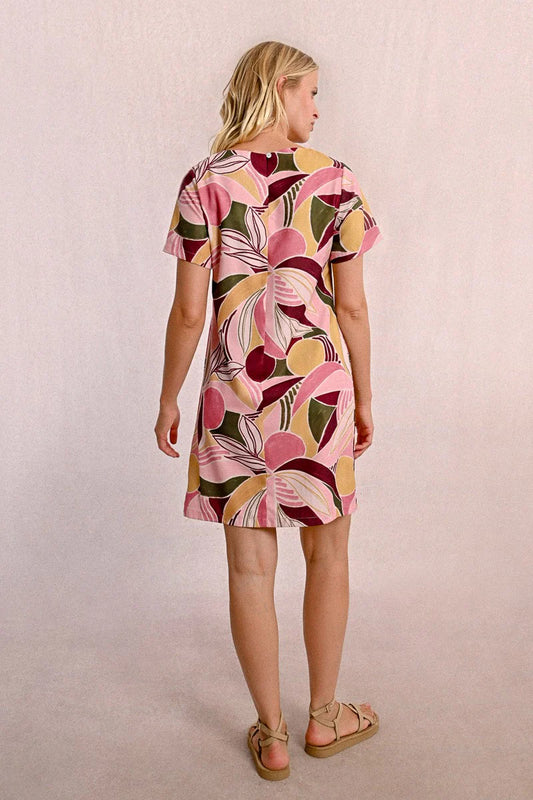 A woman in a Molly Bracken Floral Print T Shirt Dress, featuring a round collar, practical pockets, and short sleeves, stands against a neutral background. Her long blonde hair complements the vibrant outfit she pairs with chic sandals.