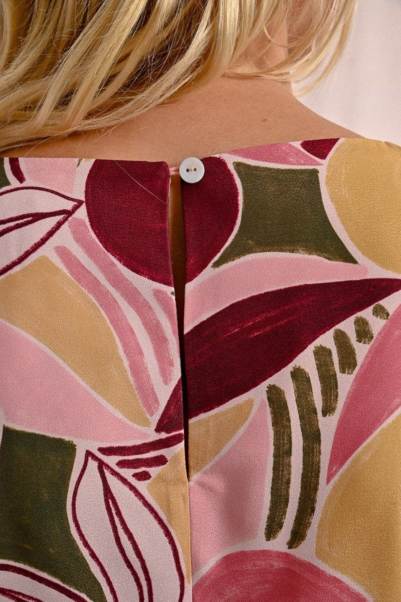 A woman in a Molly Bracken Floral Print T Shirt Dress, featuring a round collar, practical pockets, and short sleeves, stands against a neutral background. Her long blonde hair complements the vibrant outfit she pairs with chic sandals.