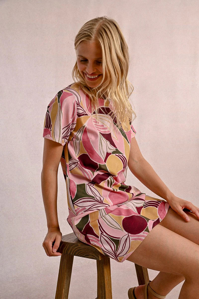 A woman in a Molly Bracken Floral Print T Shirt Dress, featuring a round collar, practical pockets, and short sleeves, stands against a neutral background. Her long blonde hair complements the vibrant outfit she pairs with chic sandals.