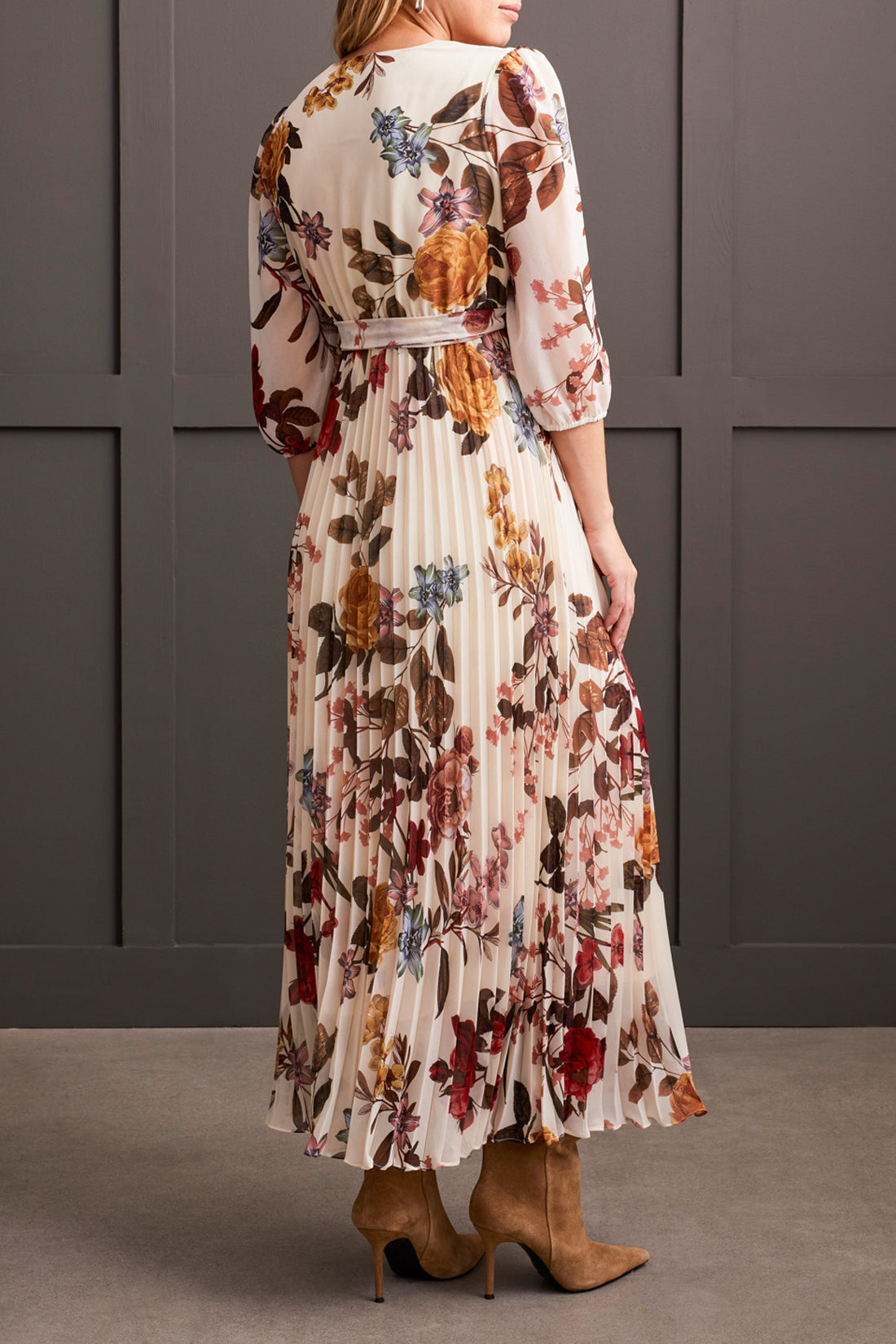 A person stands against a gray paneled background wearing Tribal's 3/4 Sleeve V Neck Dress with Side Tie, featuring floral detailing and a pleated design that accentuates the colorful flowers. The outfit is completed with tan boots.