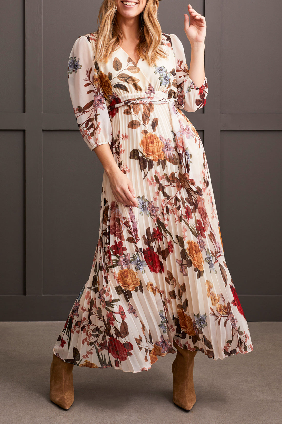 A person stands against a gray paneled background wearing Tribal's 3/4 Sleeve V Neck Dress with Side Tie, featuring floral detailing and a pleated design that accentuates the colorful flowers. The outfit is completed with tan boots.