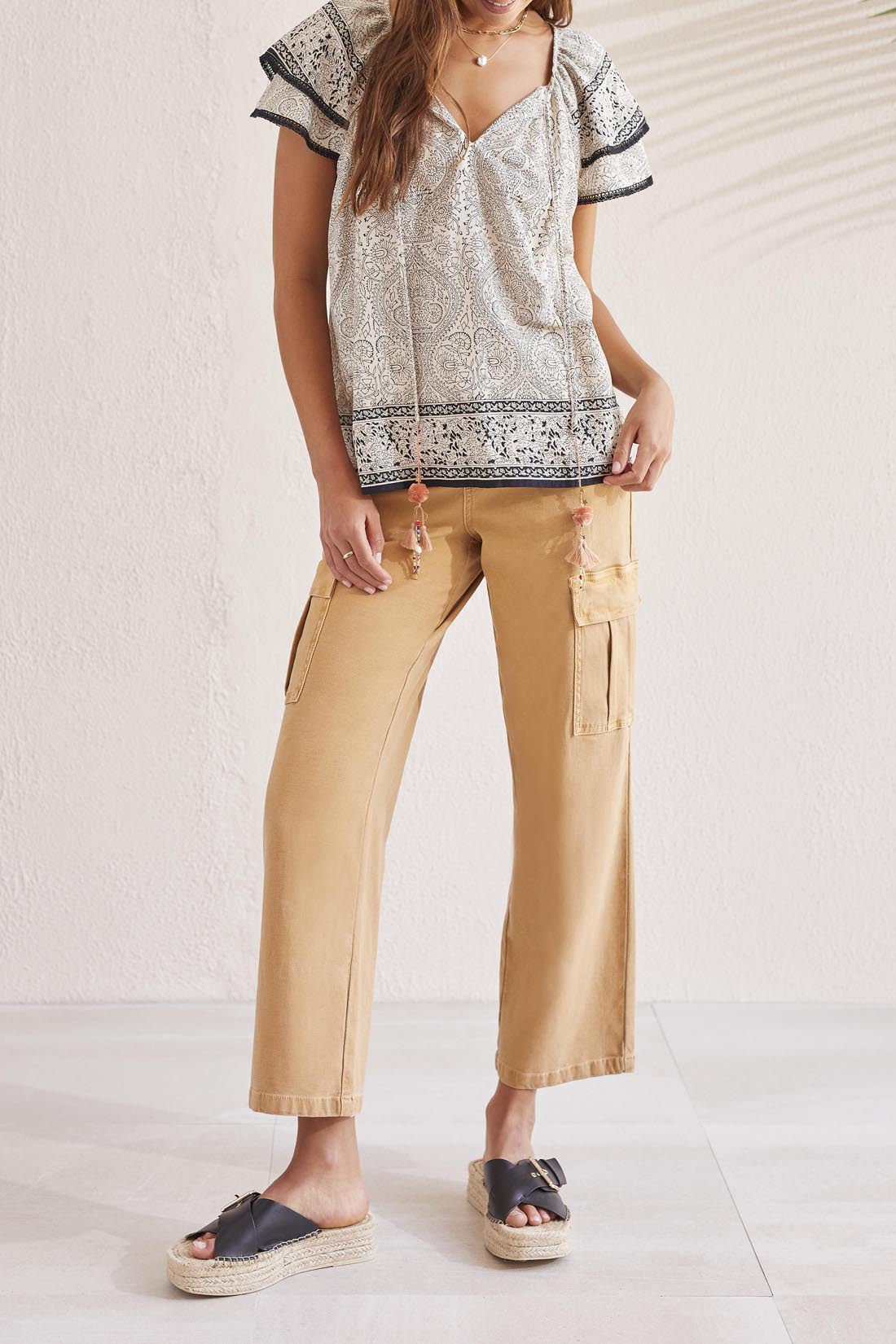 A woman facing away, wearing a Tribal Flutter Short Sleeve Blouse and beige trousers, standing against a textured wall.