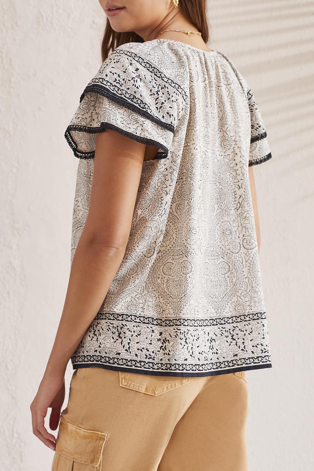 A woman facing away, wearing a Tribal Flutter Short Sleeve Blouse and beige trousers, standing against a textured wall.